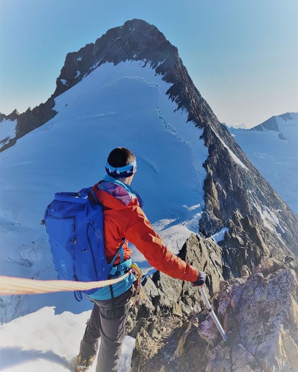 Climber with ice axe and backpack on snowy mountain ridge, wearing crampons and harness, clear sky.