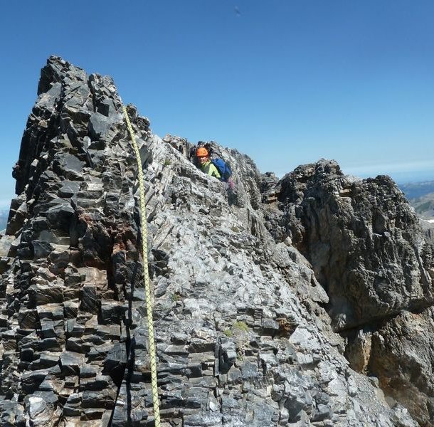 Climber with helmet and harness ascends rocky mountain ridge, secured by rope, under clear blue sky.