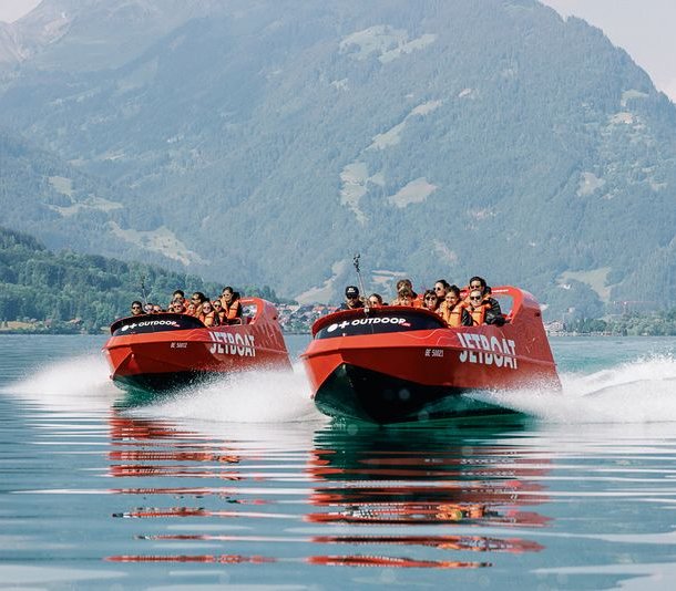 Zwei rote Jetboote mit Personen in Schwimmwesten fahren auf einem See, umgeben von Bergen.