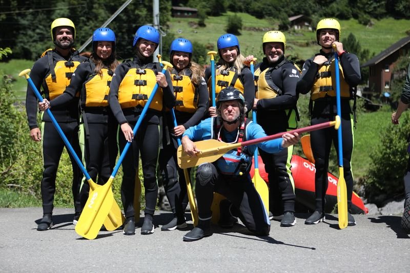 Eine Gruppe in Neoprenanzügen, Schwimmwesten und Helmen hält Paddel; im Hintergrund Berge.