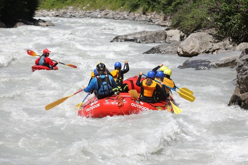 Personen in Helmen und Schwimmwesten raften in einem roten Boot auf einem Fluss in den Bergen.