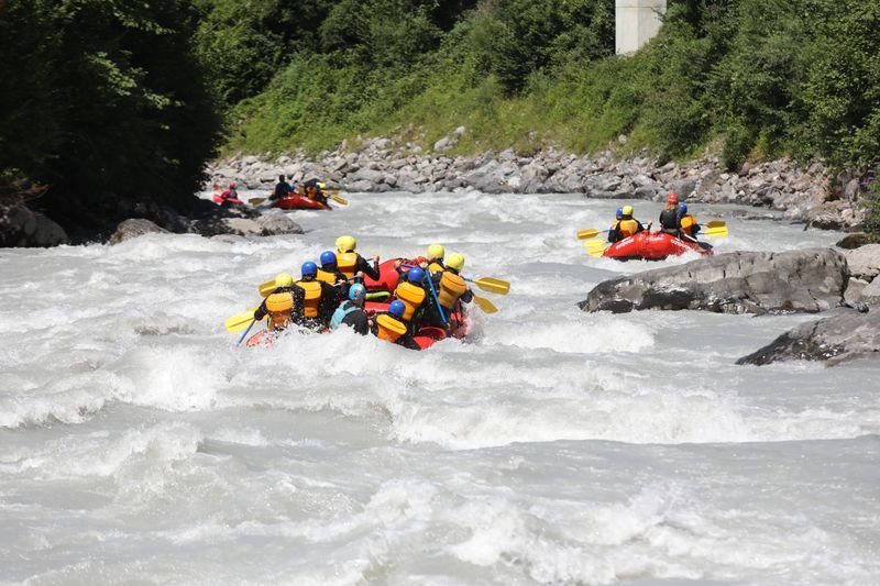 Personen in Schlauchbooten mit Helmen und Schwimmwesten beim Rafting auf einem Fluss in den Bergen.