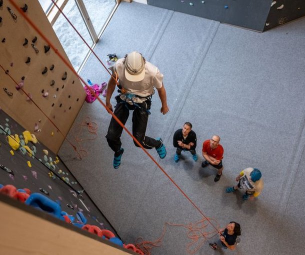 Person in climbing gear rappelling indoors, with others watching below on a padded floor.