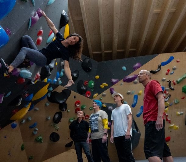 Person climbing an indoor bouldering wall with colorful holds, observed by four others in casual attire.