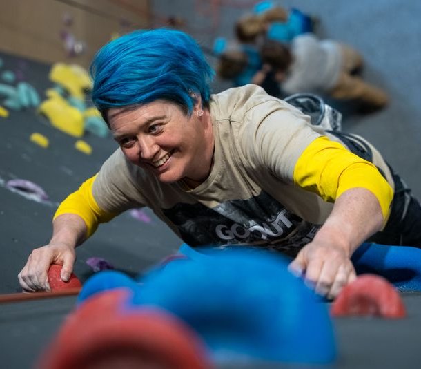 Person with blue hair climbing an indoor wall, wearing a harness and casual clothing.