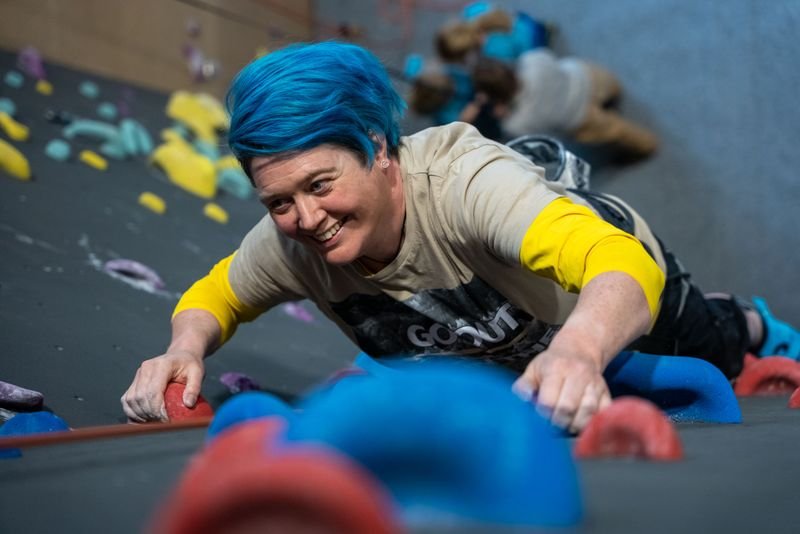 Person with blue hair climbing an indoor wall, wearing a harness and casual clothing.