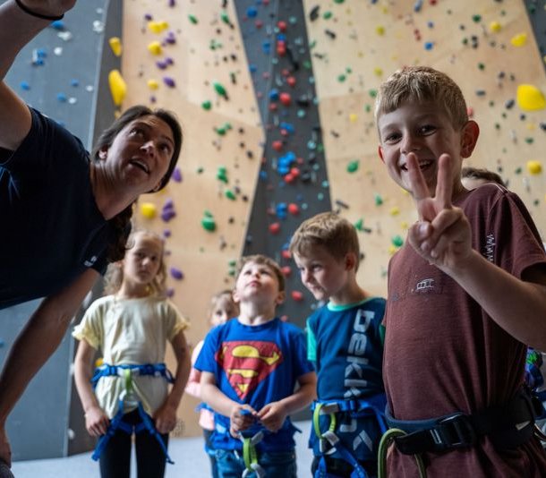 Children in harnesses listen to an instructor at an indoor climbing wall.