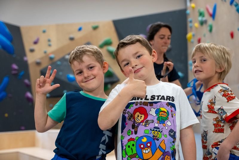 Children in a climbing gym with colorful holds on the wall; an adult is in the background.
