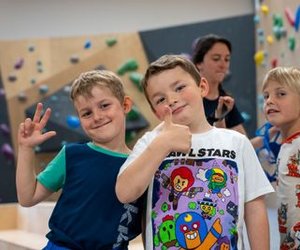 Children in a climbing gym with colorful holds on the wall; an adult is in the background.