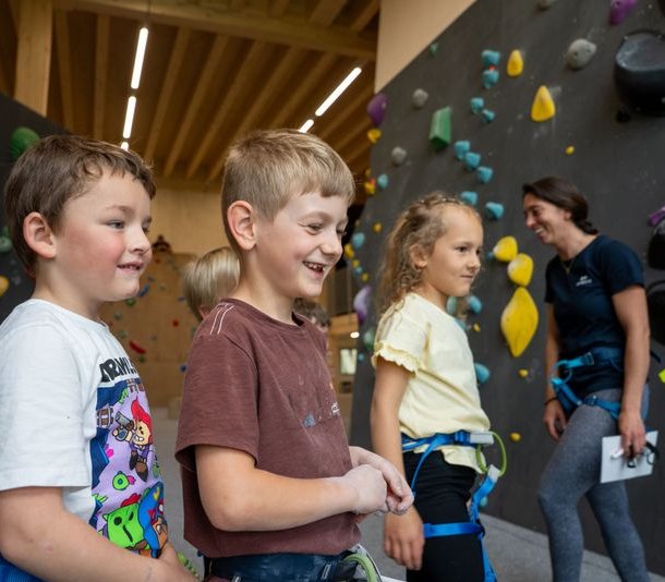Children with harnesses in an indoor climbing gym, colorful holds on walls, adult guide present.