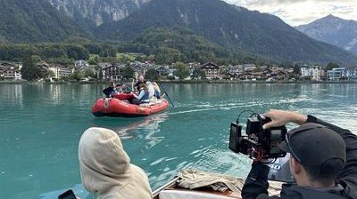 Personen in einem Schlauchboot auf einem See, umgeben von Bergen, mit Kameraaufnahmen vom Boot aus.