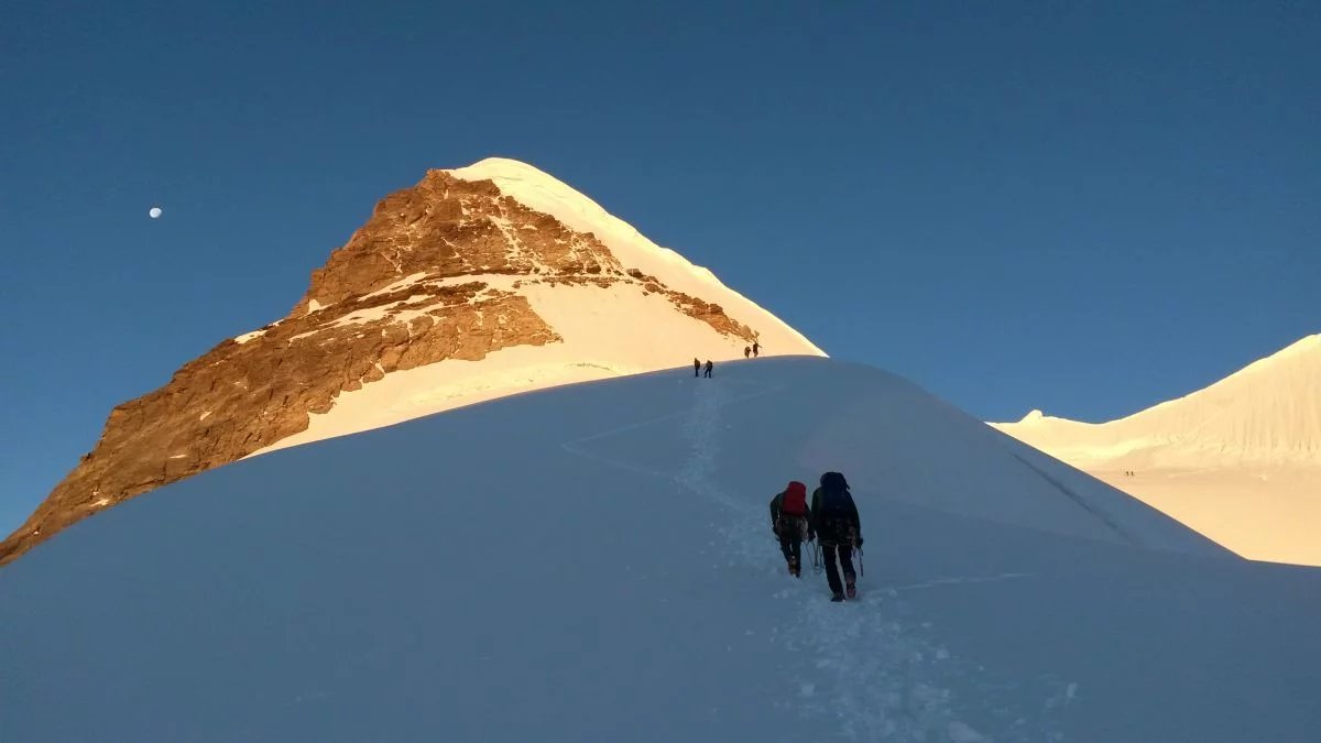 Climbers with backpacks and ropes ascend a snowy mountain slope under a clear blue sky.