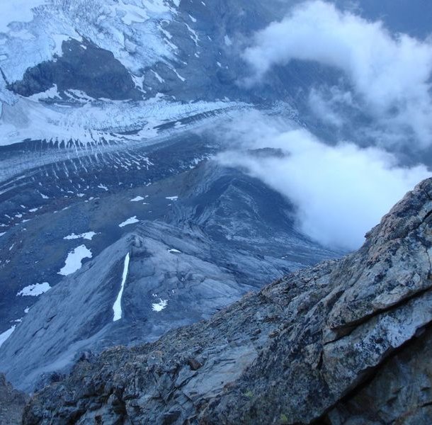 Rocky mountain peak with snow patches and clouds, overlooking a glacier and rugged terrain below.