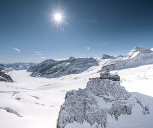Snow-covered mountains with an observatory on a rocky peak under a clear blue sky and bright sun.