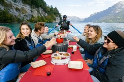 Personen in Schwimmwesten essen Fondue auf einem Boot auf einem See, umgeben von Bergen.