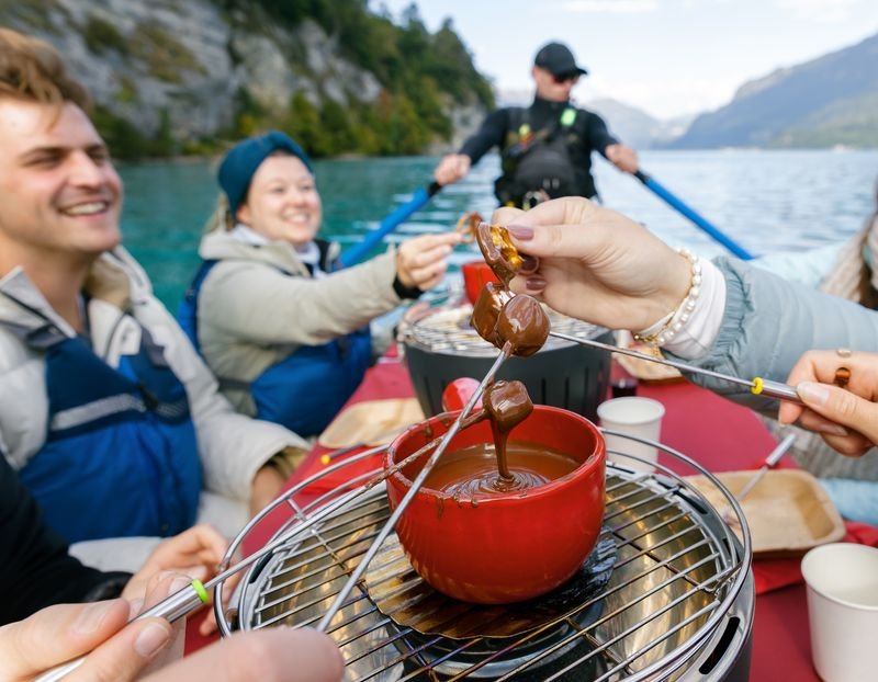 Personen in einem Boot auf einem See genießen Schokoladenfondue; sie tragen Schwimmwesten.
