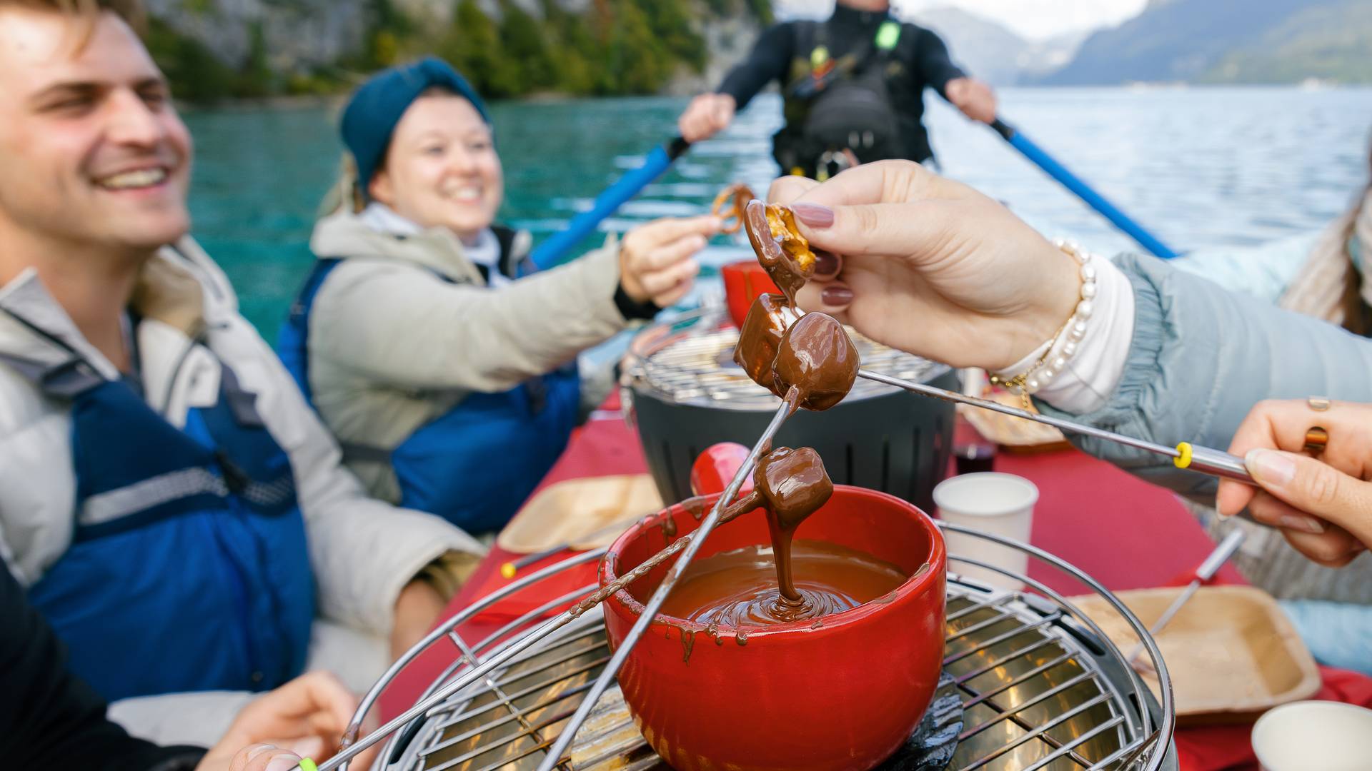 People in a boat on a lake are enjoying chocolate fondue; they are wearing life jackets.