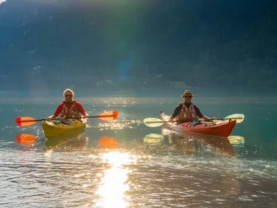 Two people wearing life jackets and hats kayaking on a calm lake with mountains in the background.