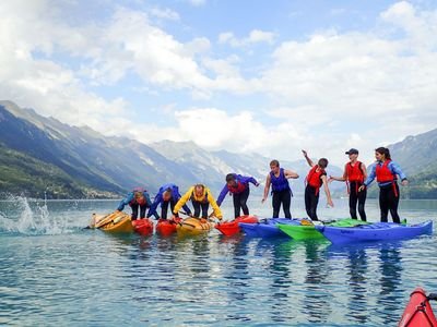 Seven people wearing life jackets are balancing and jumping on kayaks on a lake with mountains in the background.