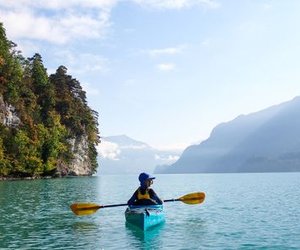 Person in blauem Kajak mit Paddel auf einem See, umgeben von Bergen und bewaldeten Klippen.