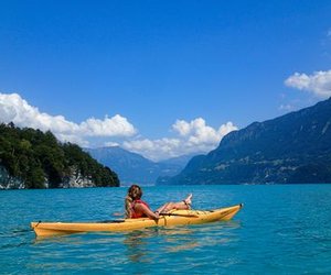 Person in gelbem Kajak auf blauem See, umgeben von Bergen und bewaldeten Ufern, unter blauem Himmel.