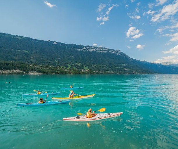 Personen in Kajaks paddeln auf einem türkisfarbenen See, umgeben von Bergen und blauem Himmel.