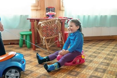 A child is sitting on a toy car in the room while being pulled by another toy car.