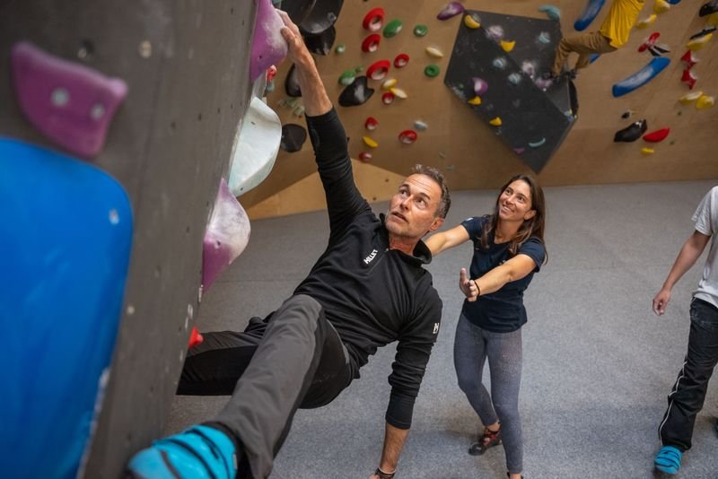 Kletterer in Kletterhalle an bunter Wand, mit Kletterschuhen und Begleiterin im Hintergrund.