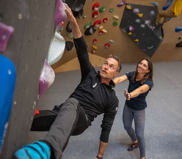 Climber in climbing gym on colorful wall, with climbing shoes and companion in the background.