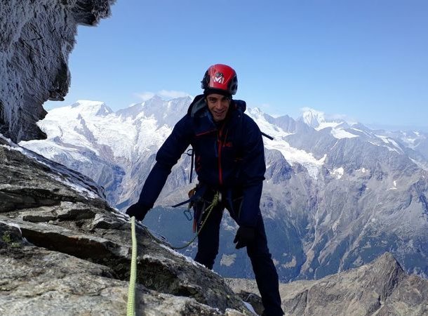 Climber with helmet and harness scaling a rocky mountain with snowy peaks in the background.