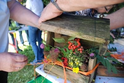 Personen dekorieren ein hölzernes Vogelhaus mit bunten Blumen und Beeren auf einem Tisch im Freien.