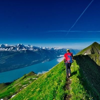 Eine Person wandert auf einem schmalen - mit Gras bewachsenen - Grat. Am Horizont ist eine Bergkette zu sehen und unten im Tal ein großer See.