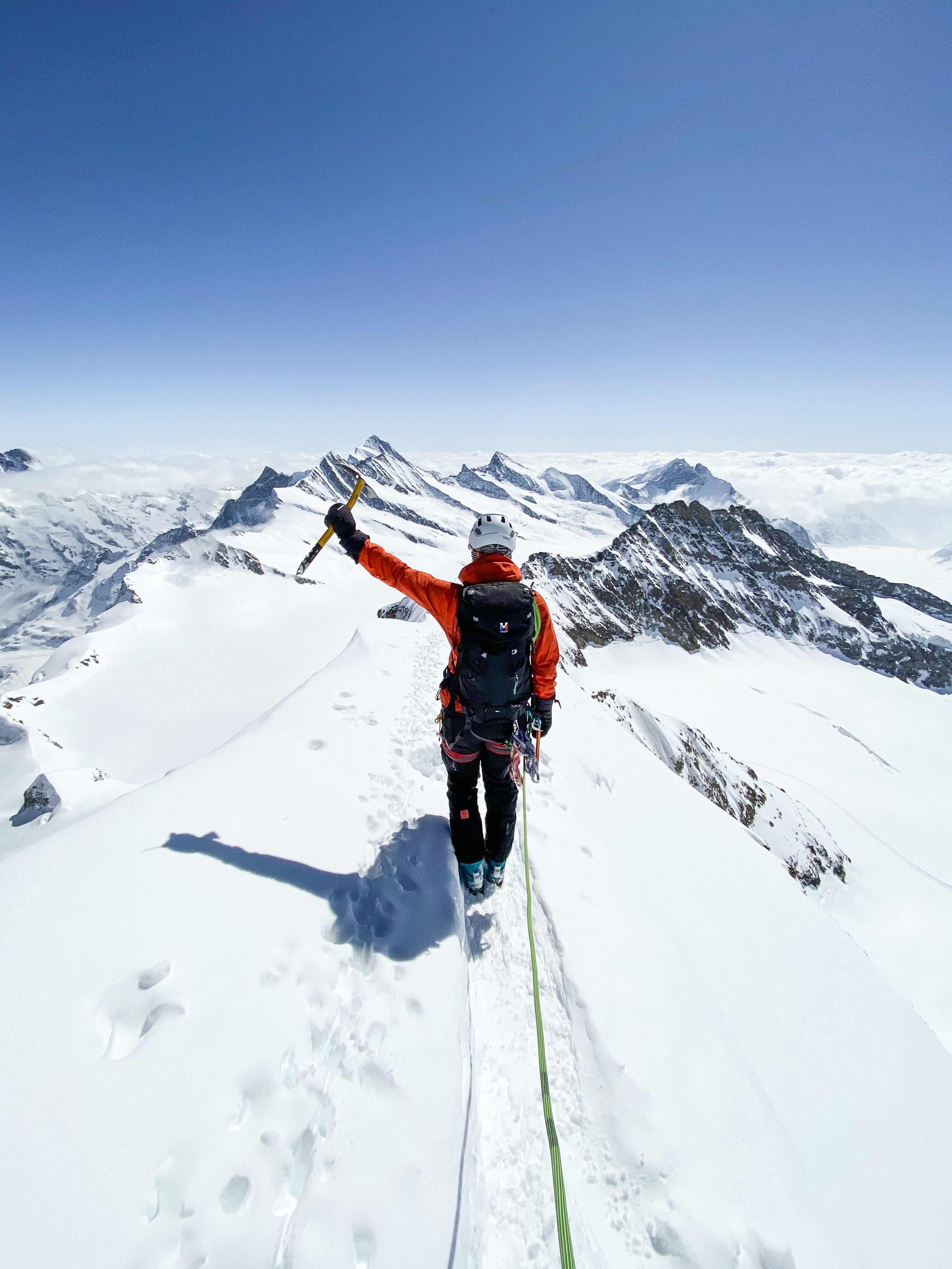 Climber on snowy mountain ridge, wearing helmet and harness, holding ice axe, with rope attached.