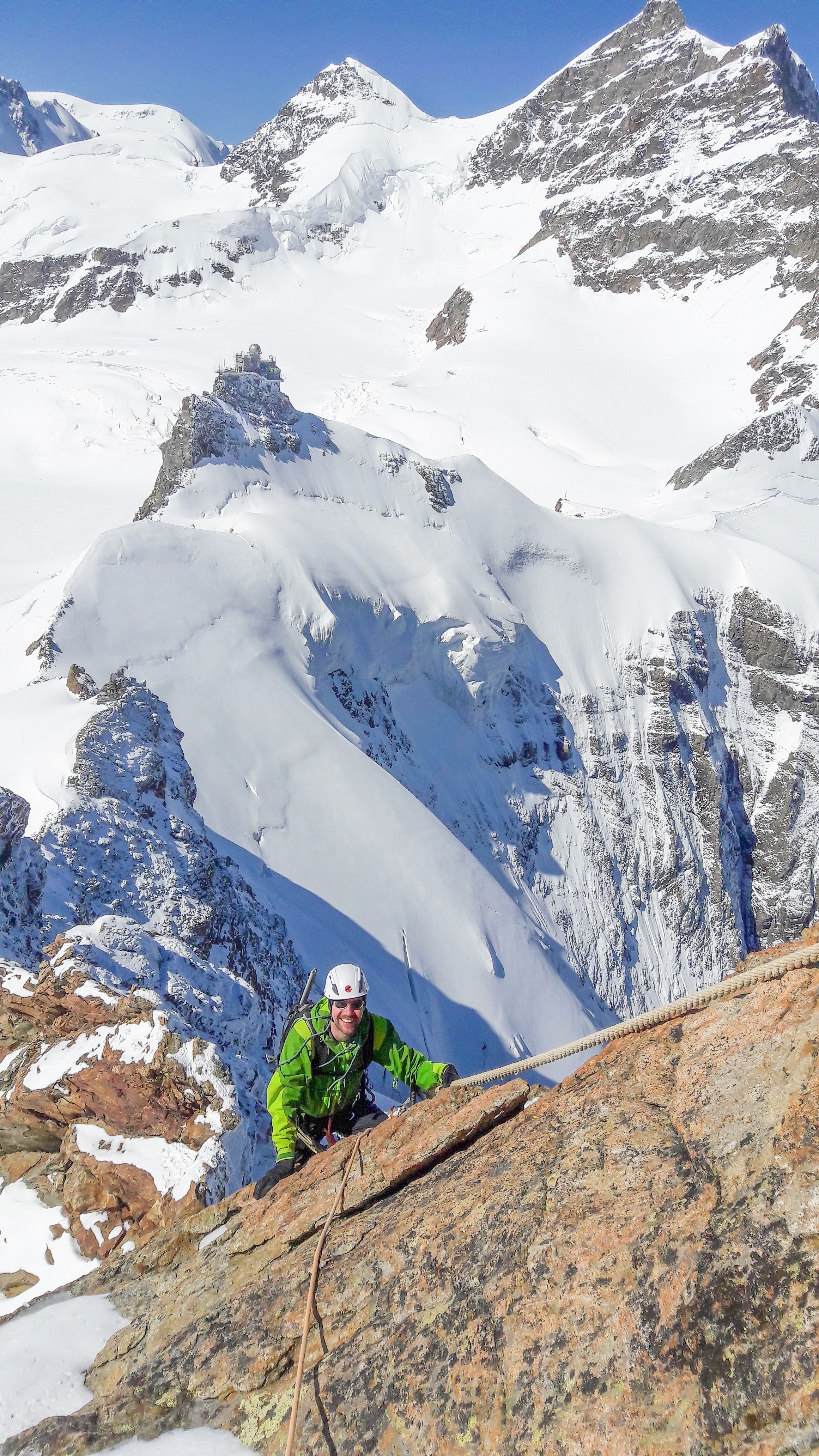 Climber in green jacket with helmet and rope ascends snowy mountain ridge with peaks in background.