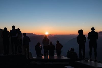 Menschen stehen bei Sonnenuntergang auf einem Bergplateau, Silhouetten vor Berglandschaft.