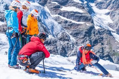 Personen in Kletterausrüstung mit Helmen und Seilen auf schneebedecktem Berg vor Felskulisse.