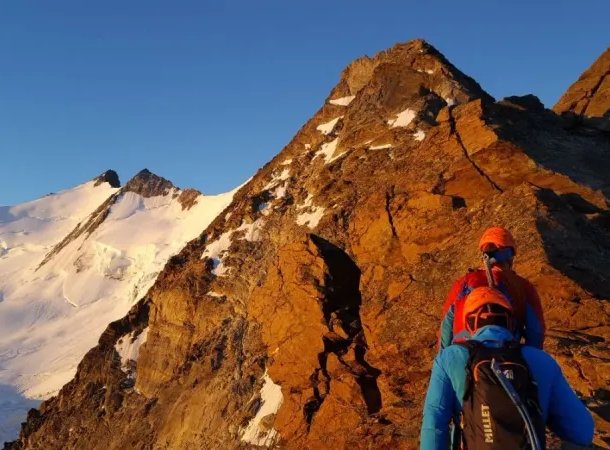Two climbers in helmets and harnesses ascend a rocky mountain ridge with snow-covered peaks nearby.