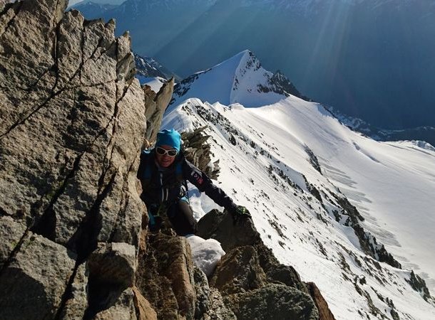 Climber in blue helmet and sunglasses ascends rocky ridge with snowy mountain backdrop.
