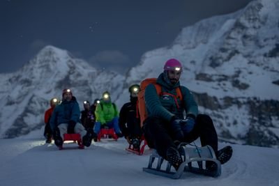 People on sleds wearing headlamps and winter gear on a snowy mountain at dusk.