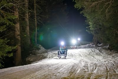 People sledding at night on a snowy forest trail, wearing helmets with headlamps.