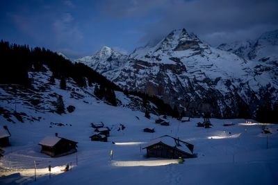 Snowy mountain village at dusk with lit cabins and towering peaks in the background.