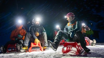 People on sleds wearing headlamps and winter gear, sitting on snow in a mountainous area at night.