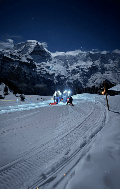 Menschen mit Schlitten und Stirnlampen auf einem verschneiten Bergpfad in der Nacht, mit Bergen im Hintergrund.