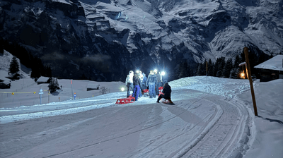 Menschen mit Schlitten und Stirnlampen auf einem verschneiten Bergpfad in der Nacht, mit Bergen im Hintergrund.
