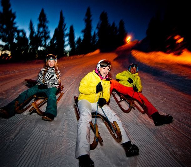 Three people are sledding on a snow slope at dusk, wearing helmets and winter clothing.