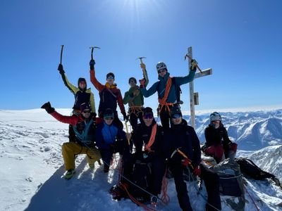 Group of mountaineers with helmets and ropes on a snow-covered summit with a mountain panorama in the background.