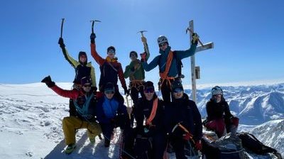 Gruppe von Bergsteigern mit Helmen und Seilen auf schneebedecktem Gipfel mit Bergpanorama im Hintergrund.