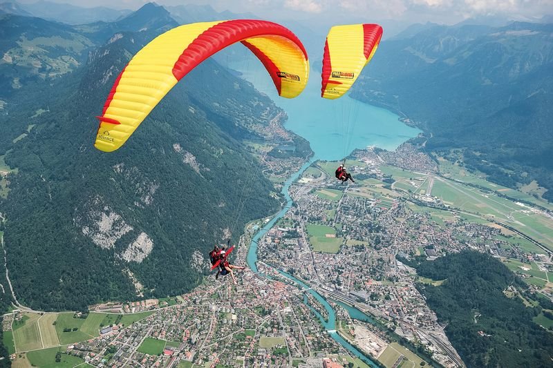 Zwei Personen beim Paragliding über einer Stadt, mit gelb-roten Schirmen und Sicherheitsgurten.