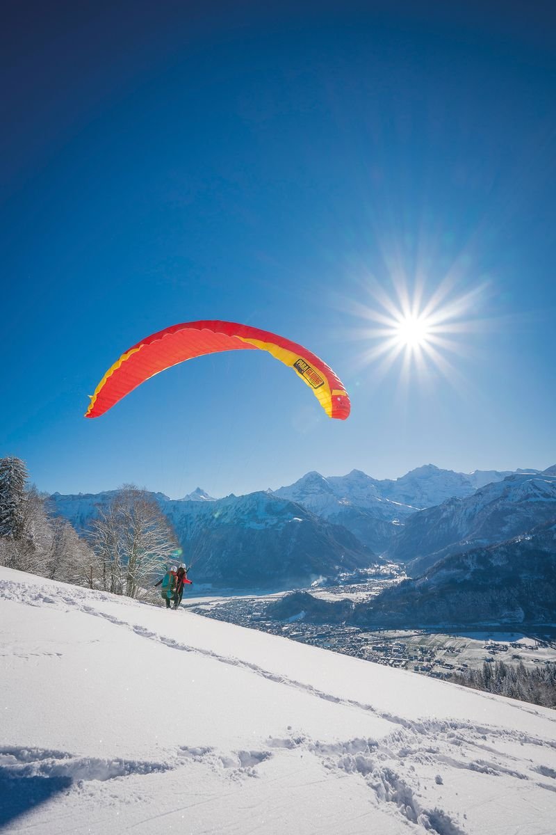 Person mit rotem Gleitschirm auf schneebedecktem Hang, Berge im Hintergrund, sonniger Himmel.