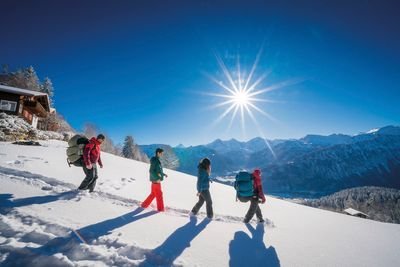Vier Personen wandern mit Rucksäcken im Schnee vor alpiner Bergkulisse bei Sonnenschein.